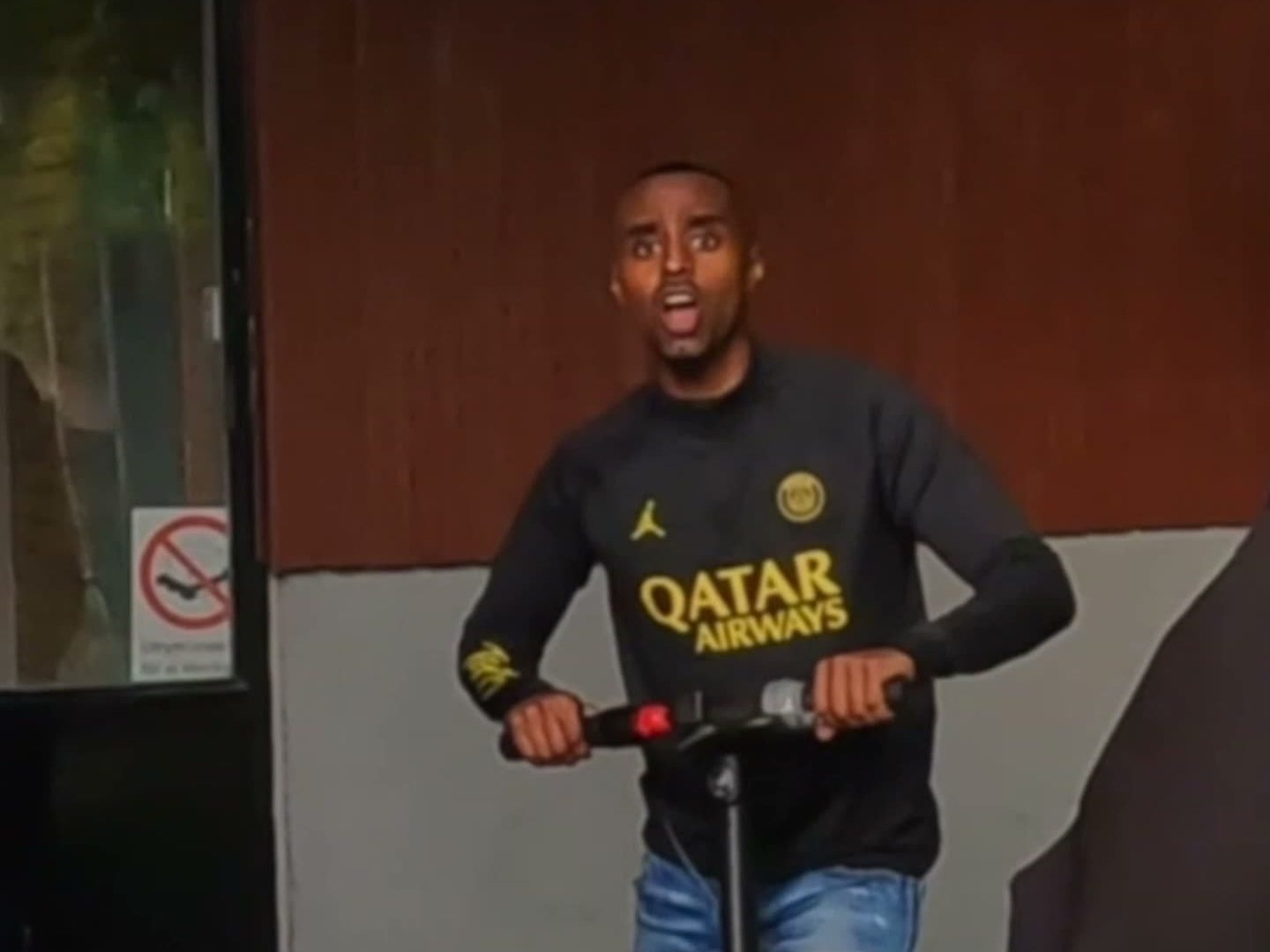 Kille i fotbollströja på elscooter framför en röd vägg, bildruta ur Goated Poesi